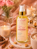 A bottle of Candi Shop Cinnamon Roll Luxury Body Oil stands on a marble board in a cozy, warm setting with candles, pink roses, cinnamon rolls, and a hot coffee cup.
