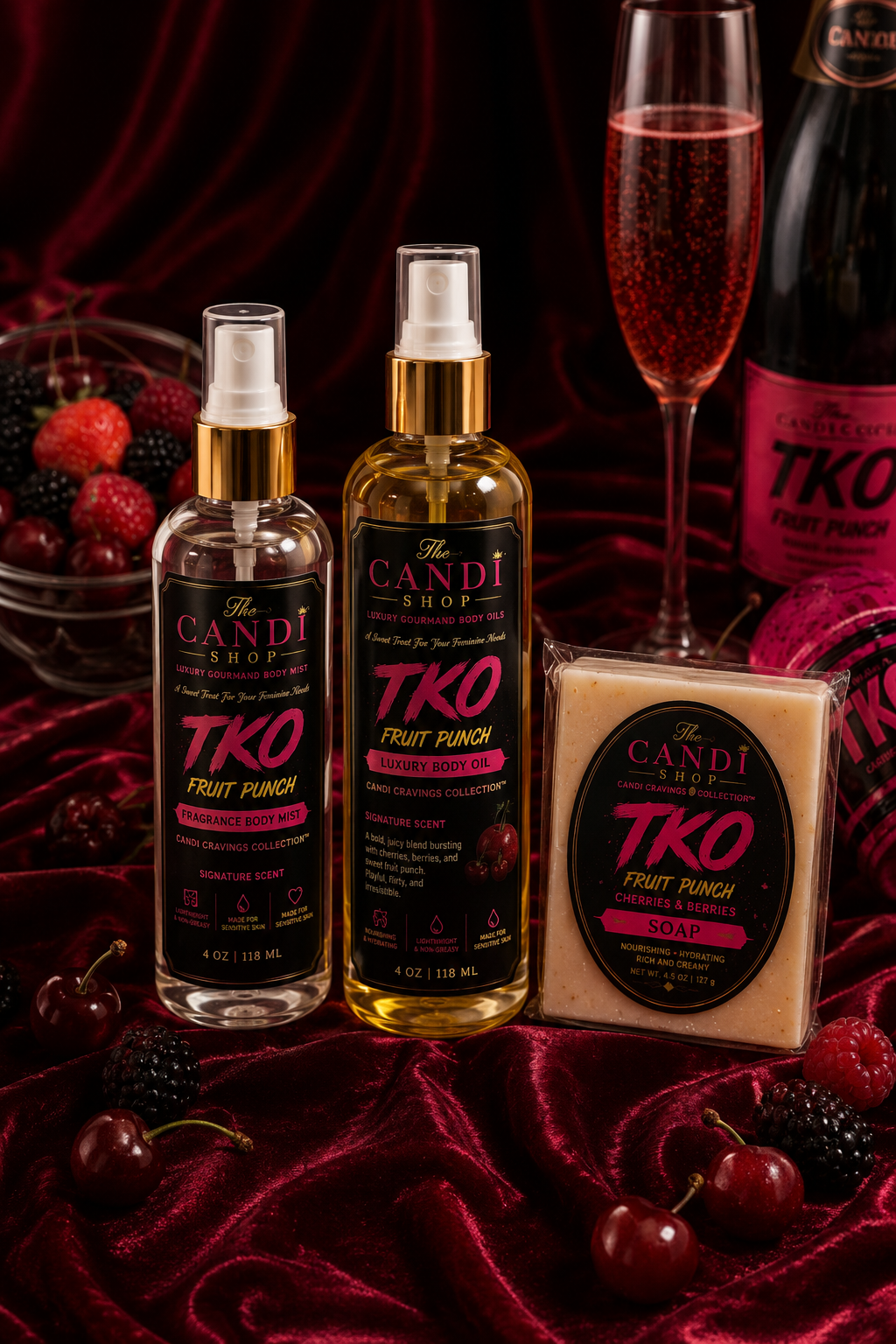 A styled flat-lay of CANDI TKO Fruit Punch flavor products on a shiny dark red satin background, including a spray bottle, a body wash bottle, a packaged fruit punch block, a glass of pink drink, and a bowl of cherries and berries.