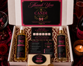 A curated gift set from Candi Shop displayed in an open white box, featuring two TKO Fruit Punch spray bottles, a reward card, an instruction card, a bar of soap, and a large thank-you card. The box is styled on a dark red fabric with rose petals, a bottle of champagne, and a lit candle nearby.
