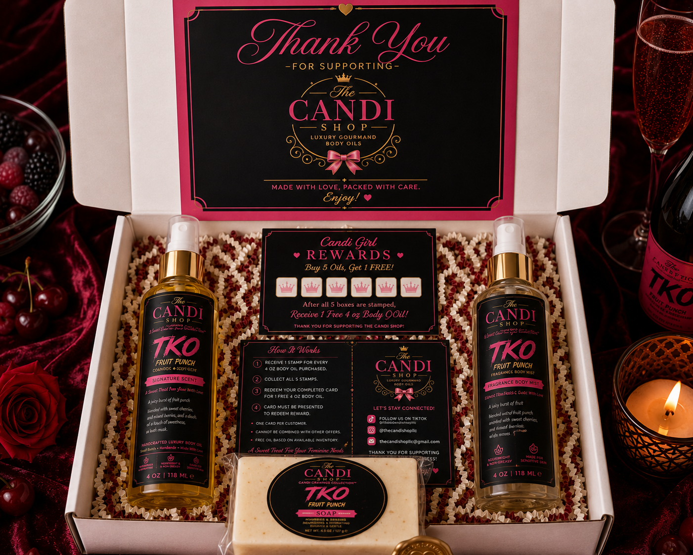 A curated gift set from Candi Shop displayed in an open white box, featuring two TKO Fruit Punch spray bottles, a reward card, an instruction card, a bar of soap, and a large thank-you card. The box is styled on a dark red fabric with rose petals, a bottle of champagne, and a lit candle nearby.