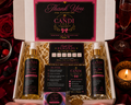 An open gift box from Candi Shop containing two bottles of 'One Night Passion' body wash, a rewards card, an instruction card, and a small packaged item, all arranged on a red velvet surface with rose petals, a champagne flute, and a lit candle.