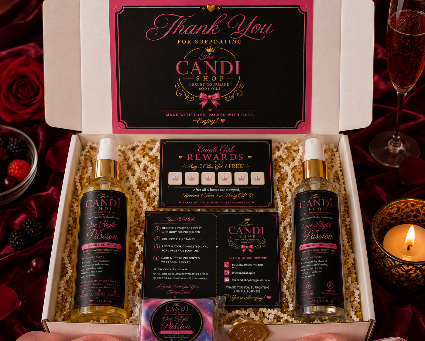 An open gift box from Candi Shop containing two bottles of 'One Night Passion' body wash, a rewards card, an instruction card, and a small packaged item, all arranged on a red velvet surface with rose petals, a champagne flute, and a lit candle.