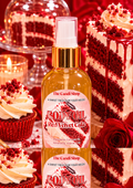 A bottle of 'The Candi Shop' Red Velvet Cake body oil is centered, surrounded by red velvet cake slices, cupcakes, and a red rose on a red background.
