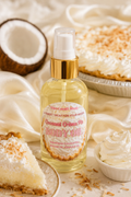 A bottle of coconut cream pie body oil by TheCandleShop.com, styled with a slice of pie, a whole coconut, whipped cream, and a pie crust on a draped cream fabric background.