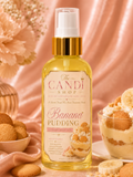 A bottle of The Candi Shop Banana Pudding Sweet & Sour essential oil mist, filled with yellow liquid and featuring a gold collar, set against a soft peach fabric backdrop with banana, cookies, and dessert props.