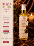 A bottle of The Candi Shop Brownie Batter Body Oil is shown against a warm, chocolate-themed background with pinecones, chocolate pieces, and a lit candle. The left side features a vertical informational panel detailing the scent notes of the body oil.