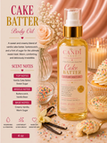 A bottle of The Candi Shop 'Cake Batter' Luxury Body Oil is displayed in a styled studio setting with cupcakes, vanilla beans, and a warm pink satin background. The left side features a text panel detailing the scent notes of vanilla cake batter, buttercups, and vanilla bean.