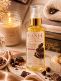 A bottle of The Candi Shop Chocolate Puffs body oil stands on a marble tray surrounded by chocolate pieces, dried flowers, a lit candle, and a folded towel. The bottle has a gold pump and clear cap, with a cream label showing chocolate imagery.
