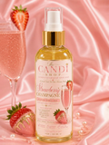 A bottle of The Candi Shop Strawberry Champagne body oil centered on a pink satin background, with a champagne flute, strawberries, and pearls as styled props.