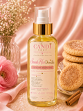 A bottle of 'The Candi Shop' 'Sneak Her Doodle' Luxury Body Oil, styled with pink satin fabric, baby's breath flowers, cinnamon sticks, and sugar cookies on a light beige surface.