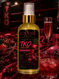 A bottle of The Candi Shop TKO Fruit Punch Luxuriously Body Oil, filled with golden-yellow oil and featuring a black label with pink and gold text, styled on a dark red fabric with champagne glasses, strawberries, and a bottle in the background.